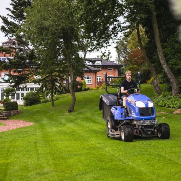 Person riding a blue lawn mower, mowing a well-kept grassy yard with trees and a brick house in the background