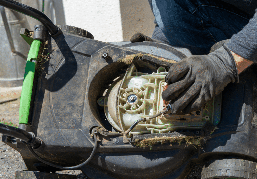 Gloved hands work on a lawn mower with its cover removed, exposing the engine and internal parts outside.